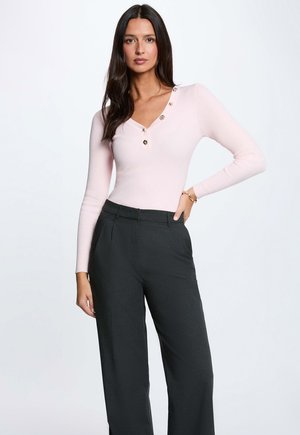 Donna con i capelli lunghi e scuri che indossa una maglia rosa chiaro a maniche lunghe con bottoni e pantaloni neri a vita alta con gambe larghe, in piedi con una mano sul fianco.