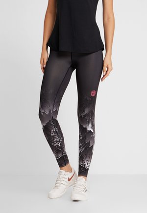 Kobieta nosząca czarne legginsy sportowe z szarym abstrakcyjnym wzorem i różowym logo na udzie, w połączeniu z białymi trampkami i czarnym topem.