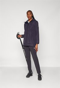 Giacca impermeabile blu navy con cappuccio, chiusura a zip e tasche, abbinata a pantaloni scuri e scarpe da trekking nere, mentre tiene un bastone da trekking.