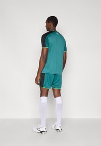 Tirkīzzila sporta krekls un šorti ar melnām detaļām, zaļu apdari un baltām zeķēm. Audums šķiet viegls, piemērots sportiskām aktivitātēm.