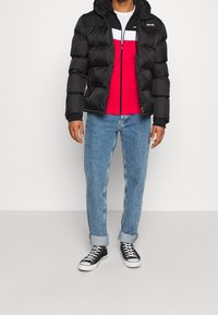 Černá péřová bunda s kapucí, pod ní červená a bílá mikina na zip, světle modré džíny a černé tenisky nad kotníky.