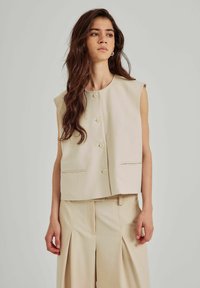 Gilet beige senza maniche con scollatura rotonda, chiusura frontale con bottoni e tasche laterali; abbinato a pantaloni plissettati beige chiaro abbinati.