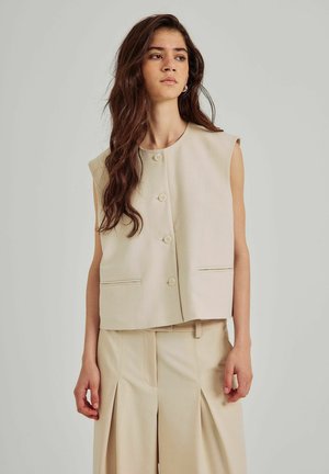 Beige mouwloze vest met ronde halslijn, knoopsluiting aan de voorkant en zijzakken; gecombineerd met bijpassende lichtbeige geplooide broeken.
