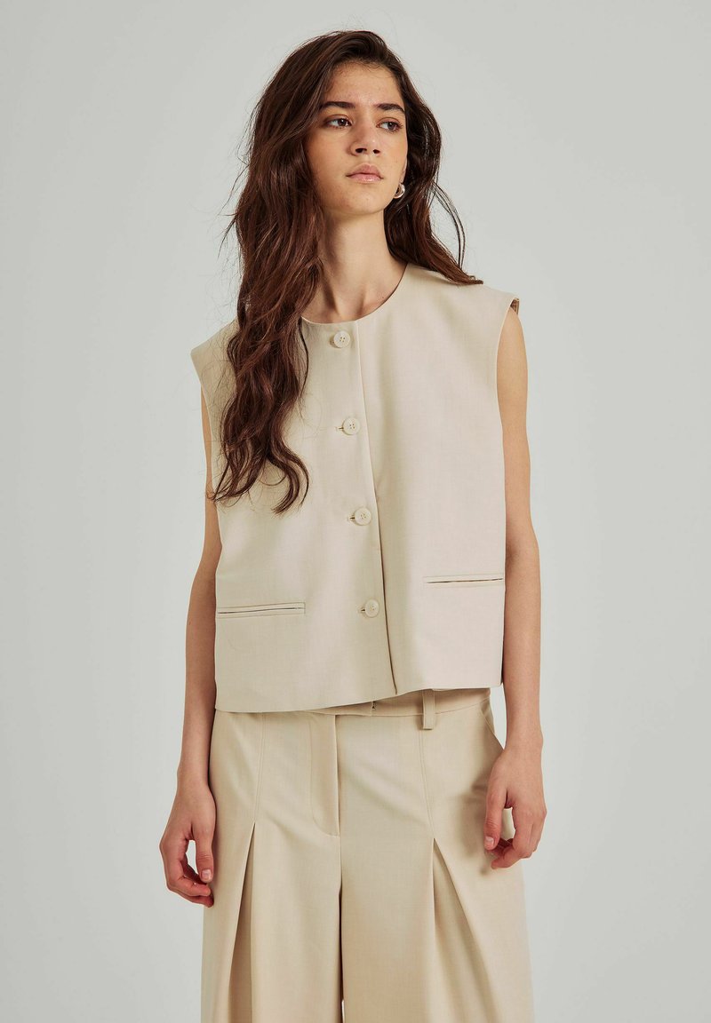 Gilet beige senza maniche con scollatura rotonda, chiusura frontale con bottoni e tasche laterali; abbinato a pantaloni plissettati beige chiaro abbinati.