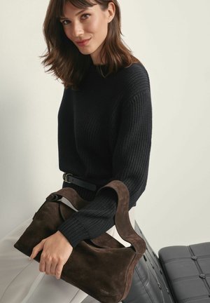 Brązowa torebka z zamszu o miękkim, zaokrąglonym kształcie i szerokim pasku, trzymana przez osobę ubraną w czarny sweter ze wzorem prążków na tle w neutralnych kolorach.