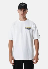 Maglietta bianca in cotone con maniche corte, con la scritta "NEW YORK" in giallo e nero sul lato sinistro del petto. Indossata con un cappellino nero.