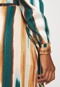 Camicia in satin con maniche lunghe e arricciate, caratterizzata da strisce verticali in teal, oro e crema. Il polsino presenta un dettaglio con bottone dorato.