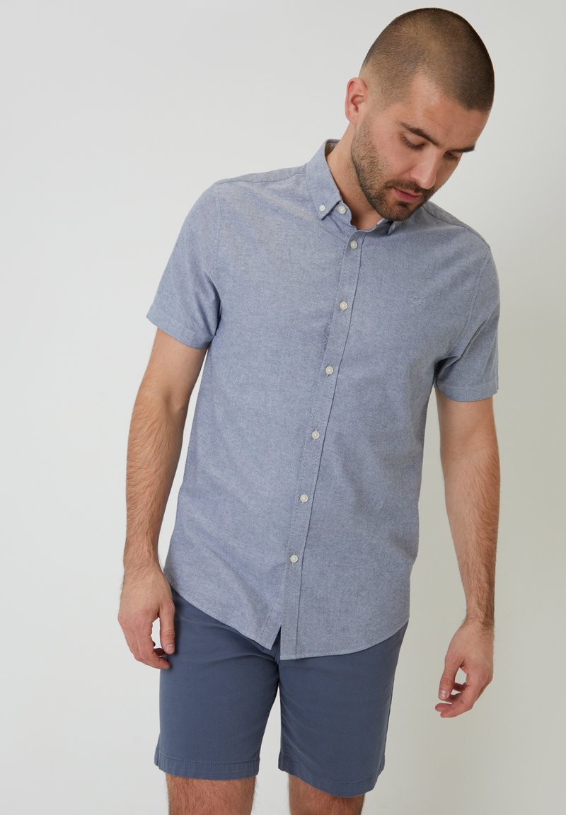 Camisa de botones de manga corta en tela azul claro, con cuello y textura suave. Combinada con pantalones cortos azul oscuro.