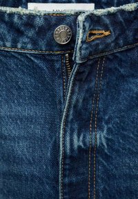Blaue Jeans aus Denim verfügen über einen Reißverschluss, Kupferbeschläge und kontrastierende orangefarbene Nähte. Der Stoff zeigt eine leicht verblasste Textur.