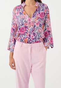 Blouse florale multicolore avec col en V et manches cloche trois-quarts, associée à un pantalon rose clair. Tissu léger et texturé.