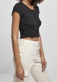 Svart cropped ribbad topp med korta volangärmar, med rund halsringning och sex knappar fram. Matchas med ljusa byxor.