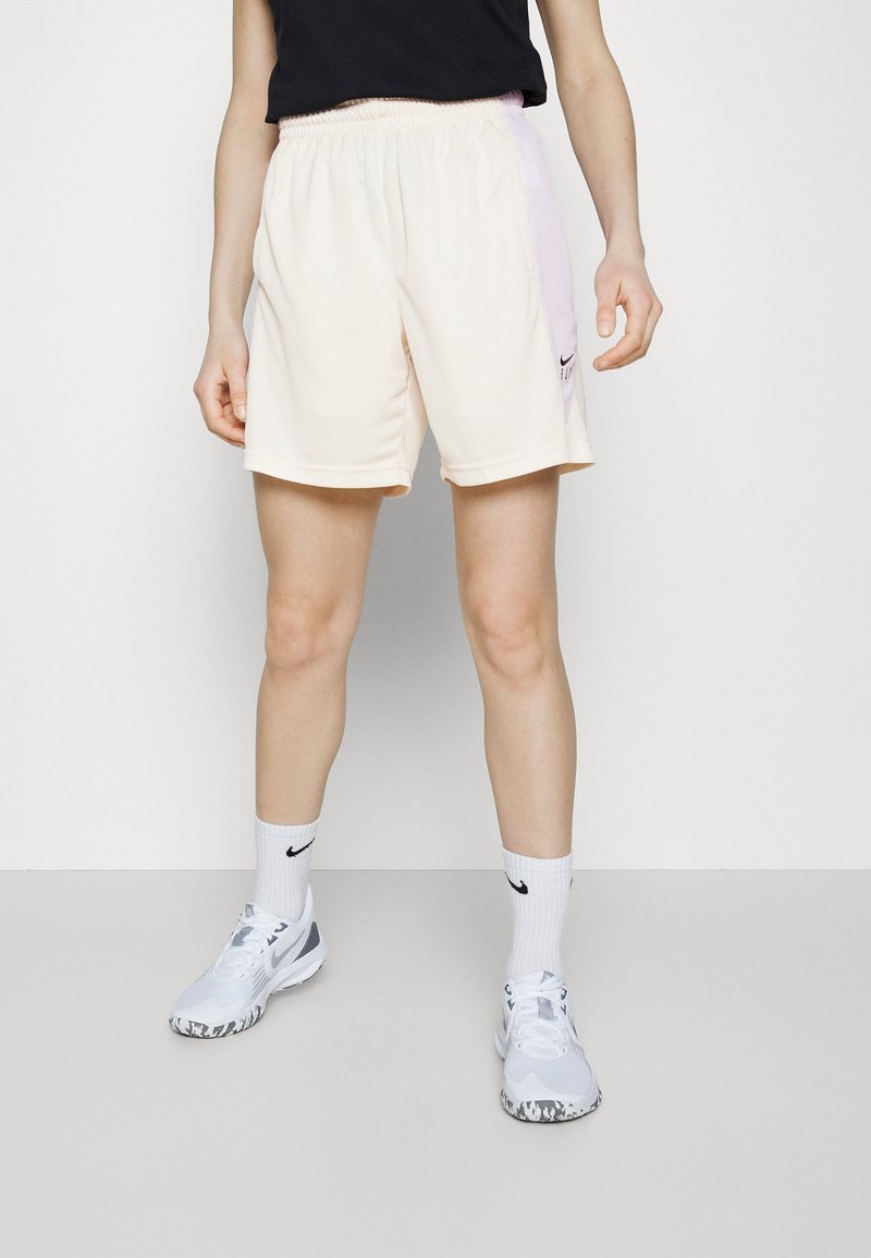 Personne portant un short de sport beige clair, des chaussettes blanches Nike et des baskets blanches Nike, debout devant un fond blanc uni.