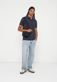 Marine polo-shirt, lyse blå jeans og hvide sneakers. Sort crossover taske med tekstureret detalje. Stående stilling mod en ensfarvet baggrund.