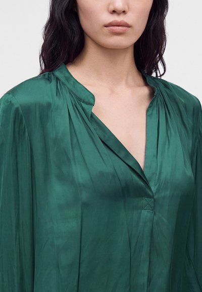 Femme aux cheveux foncés portant un chemisier en satin vert profond avec un col en V et un col froncé, montrée des épaules au menton.