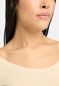 Beige off-shoulder top met een gladde textuur, voorzien van een brede halslijn en subtiele stiksels, gedragen door een persoon met vlechten.