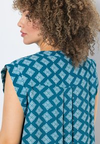 Türkise Bluse mit kurzen Ärmeln, die ein weißes Diamantmuster und Faltendetails auf der Rückseite aufweist. Glatte Stoffoberfläche.
