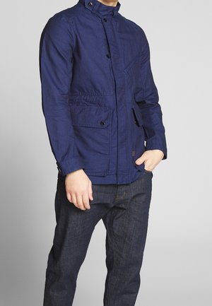 Mann trägt eine marineblaue Freizeitjacke mit Pattentaschen und dunkelblaue Jeans, steht mit einer Hand in der Tasche vor einem einfarbigen Hintergrund.