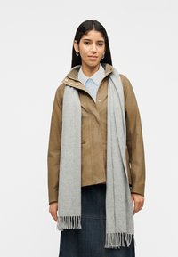 Γυναίκα με μακριά σκούρα μαλλιά, φοράει ανοιχτό καφέ μπουφάν, ανοιχτό μπλε πουκάμισο, ανοιχτό γκρι κασκόλ και σκουρόχρωμη τζιν φούστα, στέκεται μπροστά σε ένα απλό φόντο.