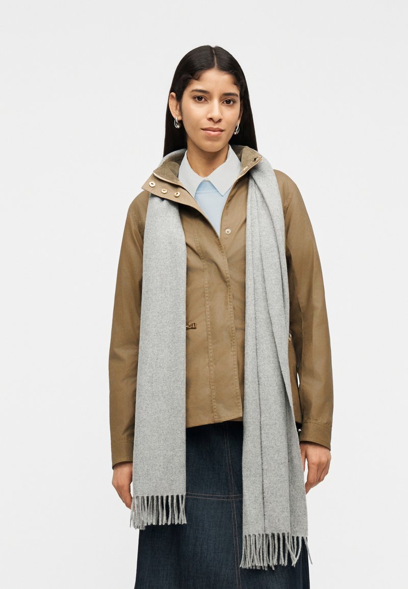 Γυναίκα με μακριά σκούρα μαλλιά, φοράει ανοιχτό καφέ μπουφάν, ανοιχτό μπλε πουκάμισο, ανοιχτό γκρι κασκόλ και σκουρόχρωμη τζιν φούστα, στέκεται μπροστά σε ένα απλό φόντο.