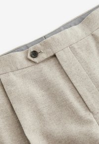 Vaaleanbeiget räätälöidyt housut, joissa on herringbone-kuvio, yksi laskos ja musta nappikiinnitys vyötäröllä. Pehmeä tekstuuri.