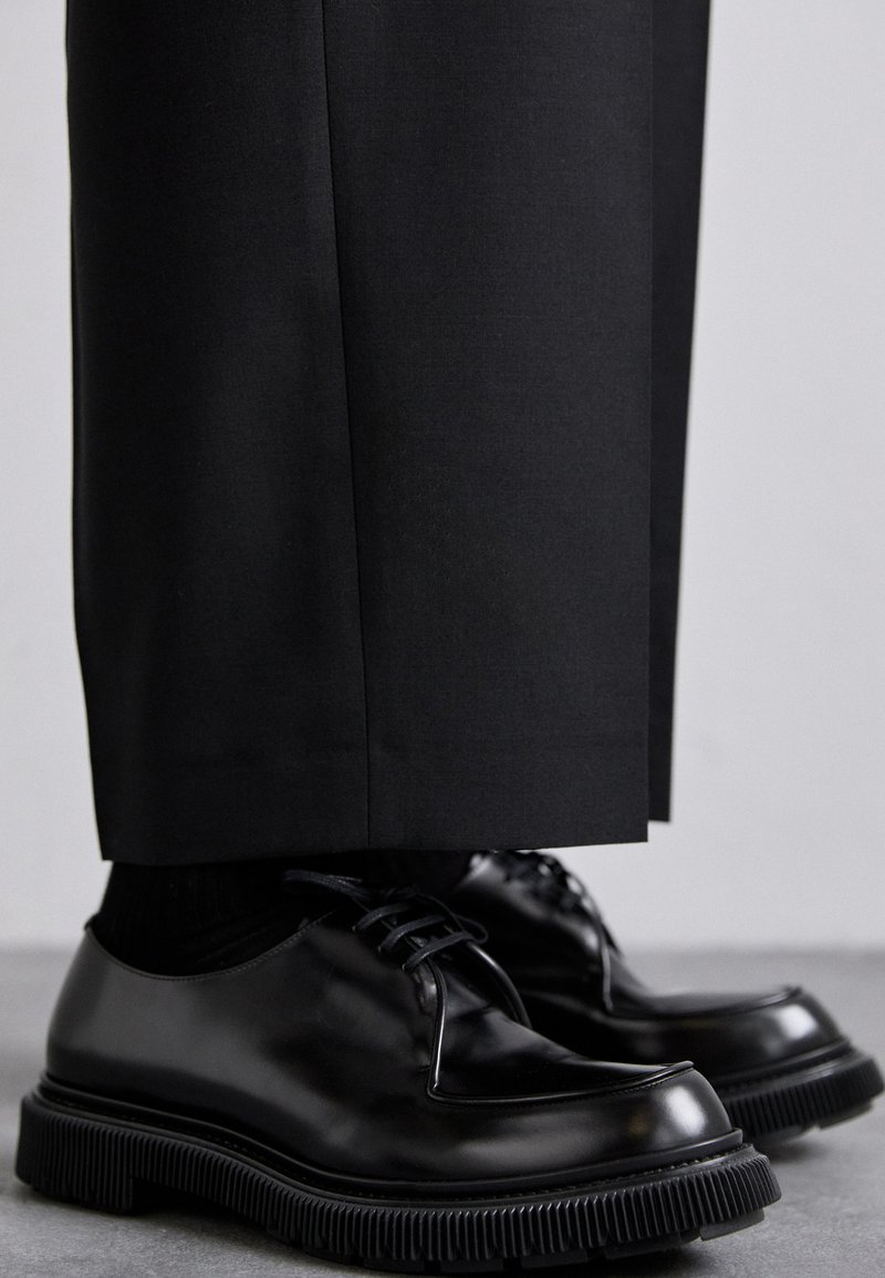 Black leather shoes with laces, featuring a rounded toe and rubber sole with ridged texture. Paired with tailored black pants.