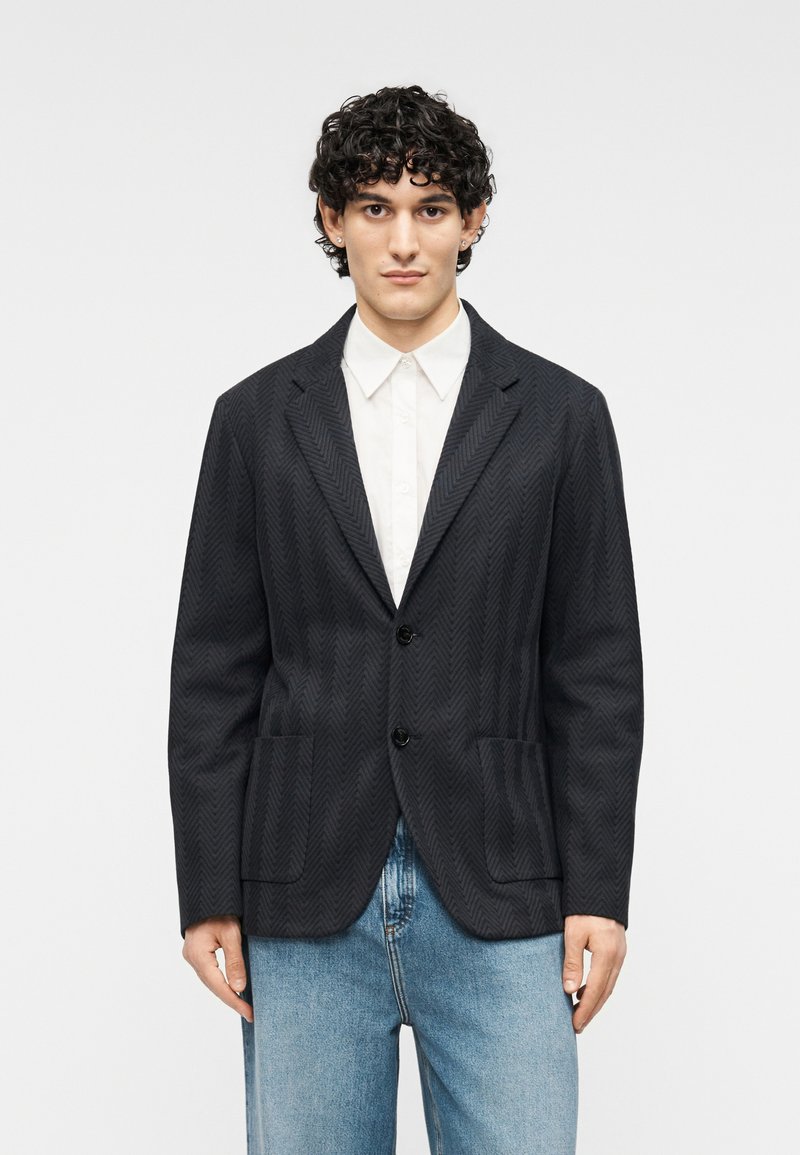 Blazer noir à motif chevrons avec deux poches avant, fermeture à un bouton, revers crantés et texture lisse. Associé à un jean bleu clair.
