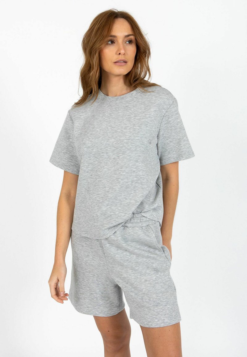 Femme aux cheveux châtain clair portant une chemise grise ample à manches courtes et un short assorti à taille élastique, debout devant un fond blanc.