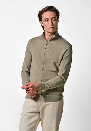 Man draagt een beige trui met rits en licht crèmekleurige broek, staat met gevouwen handen en kijkt iets naar de zijkant tegen een effen witte achtergrond.