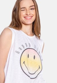 Witte mouwloze top met een grote, meerkleurige glimlachende gezichtsgrafiek en de tekst "Happy Vibes", gemaakt van lichtgewicht, gestructureerde stof.