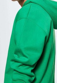 Sweat à capuche vert à la texture douce, doté d'une capuche avec cordon de serrage et de poignets côtelés, présenté de profil pour mettre en valeur le détail de la manche.