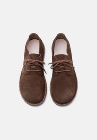Ζευγάρι καφέ σουέτ παπούτσια με κορδόνια, στρογγυλεμένες μύτες και σόλες με υφή φελλού, εμφανιζόμενα από πάνω σε λευκό φόντο.