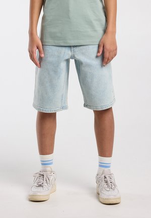 Enfant debout portant un short en denim bleu clair, des baskets blanches, des chaussettes blanches avec des rayures bleues, et un t-shirt vert, sur fond blanc.