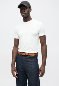 Λευκό βαμβακερό t-shirt με κοντά μανίκια, που φέρει ένα μικρό λογότυπο. Φορεμένο με σκούρα μπλε τζιν και καφέ δερμάτινη ζώνη, ολοκληρωμένο με γκρι καπέλο.