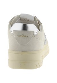 Beige suède sneaker met een crèmekleurige zool, een metallic zilveren hielstuk en het woord "victoria" gedrukt op de achterkant.