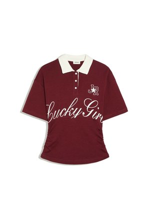 Polo manches courtes rouge foncé avec col blanc, quatre boutons, inscription "Lucky Girl" sur la poitrine et petit logo "JR" avec étoile sur le côté gauche.