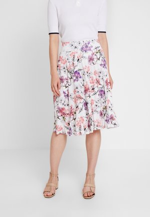 Femme portant une jupe blanche fleurie longueur genou avec des fleurs roses et violettes, des talons ouverts beiges et un haut blanc à manches courtes.