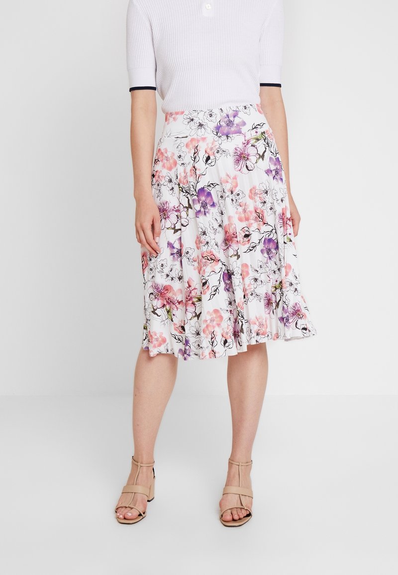 Femme portant une jupe blanche fleurie longueur genou avec des fleurs roses et violettes, des talons ouverts beiges et un haut blanc à manches courtes.
