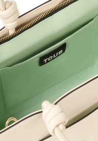 Kremowa torebka o fakturze, z zielonym wnętrzem, czarną metką z logo, złotym zamkiem i szczegółami w formie węzła na uchwycie ze sznura.