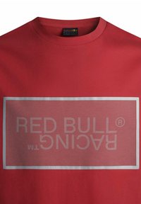 Czerwona koszulka wykonana z bawełny, z okrągłym dekoltem. Posiada duży srebrny prostokąt o siatkowanej teksturze, w którym wyraźnie widnieje napis "RED BULL RACING".