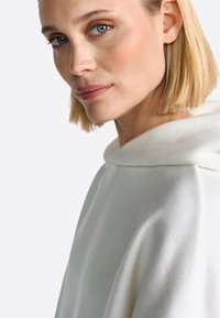 Valge kapuutsiga särk pehme tekstuuriga, laia dekolteega ja minimaalse disainiga. Kangas näeb välja sile ja kerge.