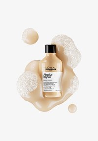 Beige shampooflaske med svart lokk, merket "L’Oréal Professionnel Absolut Repair." Omgitt av kremete bobler og væske i lys beige.