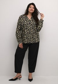 Langärmlige Bluse mit geometrischem Würfeldruck in Beige und Schwarz, kombiniert mit schwarzen Hosen. Der Stoff wirkt glatt; das Shirt hat einen V-Ausschnitt.