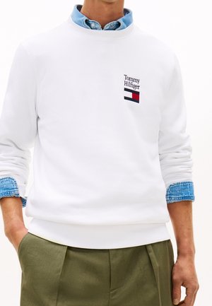 Homme portant un sweat-shirt blanc Tommy Hilfiger par-dessus une chemise en denim bleu et un pantalon vert olive, les mains dans les poches.