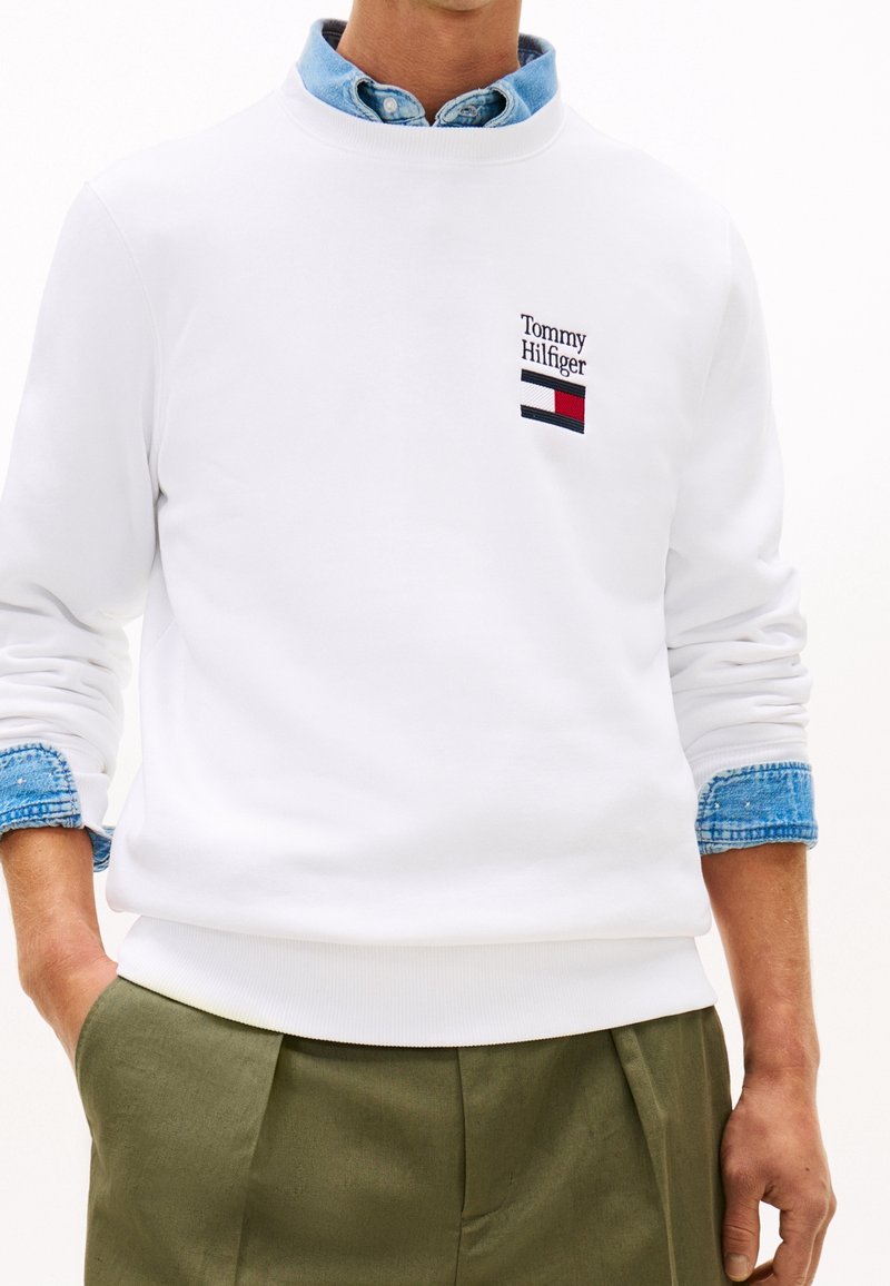 Homme portant un sweat-shirt blanc Tommy Hilfiger par-dessus une chemise en denim bleu et un pantalon vert olive, les mains dans les poches.