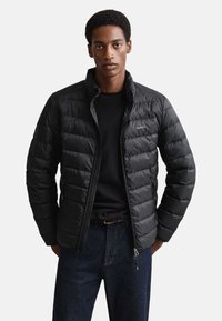 Juoda pūkinė striukė su siuvinėtomis dalimis, aukšta apykakle, priekiu su užtrauktuku ir logotipo detalėmis. Dėvima ant juodo megztinio su mėlynomis džinsinėmis kelnėmis.