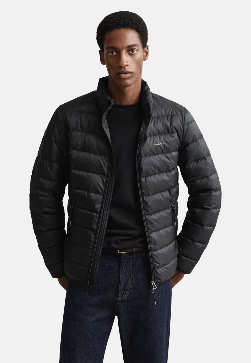 Fekete pufi dzseki, steppelt részekkel, magas gallérral, elülső cipzárral és logó részlettel. Fekete pulóver fölött viselve, kék farmernadrággal.