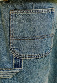 Denimist teksapüksid kandilise tagataskuga, topeltõmmeldud oranžide õmblustega ja vööaasaga, mis asub tasku all.