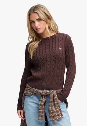 Kvinde med langt blond hår iført en brun kabelstrikket sweater, lyseblå jeans og en bundet ternet skjorte omkring taljen.