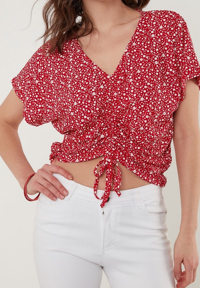 Haut floral rouge à col en V, manches courtes et taille à cordon. Associé à un jean blanc taille haute. Tissu léger avec une finition texturée.