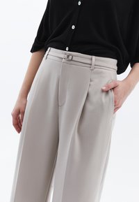 Lichtgrijze gebeitelde broek van soepel materiaal, met een rechte pijp, voorplooien en een zilveren gesp als detail.